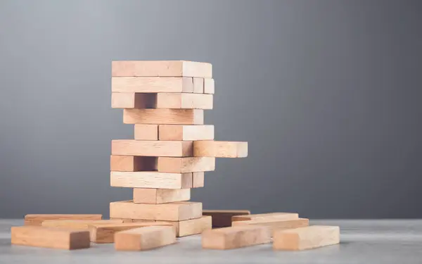 stock image Precarious stack of wooden blocks towers on the table against a grey wall background. Symbolizes risk, uncertainty, and careful decision-making, much like in a game of balancing strategy and chance