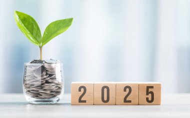 2025, paralarla dolu cam bir kavanozun yanındaki tahta bir blokta parlak bir beyaza karşı tahta bir masanın üzerine yazılmış. Finansal planlamayı, tasarrufu ve etkili para yönetimini sembolize eder