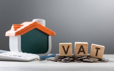 VAT is written on a wooden block, accompanied by a pile of coins and a house model, placed on a wooden table against grey background. VAT, financial management, and home-related financial planning. clipart