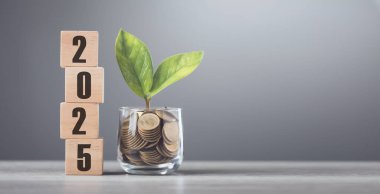 2025, paralarla dolu cam bir kavanozun yanındaki tahta bir blokta parlak bir beyaza karşı tahta bir masanın üzerine yazılmış. Finansal planlamayı, tasarrufu ve etkili para yönetimini sembolize eder