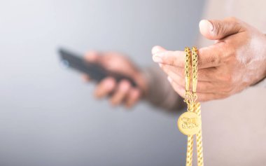 Close-up of a man holding a gold necklace in his left hand while checking his phone with his right hand. Concept of monitoring gold prices, symbolizing market speculation and investment trends clipart