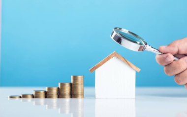 Üzerinde büyüteç tutan bir el olan bozuk para yığınının yanındaki ahşap ev modeli. Görüntü, ev bakımı ve onarım maliyetlerini analiz ve hesaplama kavramını temsil ediyor.