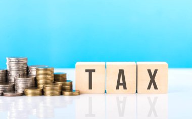 Vergi, bir yığın madeni paranın yanındaki tahta bloğa yazmaktır. Açık mavi arka plana karşı. Vergi bilinci, mali planlama, tasarruflar ve etkili para yönetimi