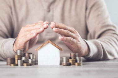 Bir iş adamı, bir yığın bozuk para ve bir ev modeline karşı koruyucu bir el hareketi yapıyor. Resim, varlık koruma kavramını temsil ediyor, mali yatırımların ve mülkiyetin korunmasını vurguluyor