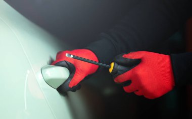 Tornavida tutan bir adamın eldivenli eli, arabanın kapı kolunu açmaya çalışıyor. Görüntü araba hırsızlığı konseptini temsil ediyor, araç güvenliğinin riskini ve önemini vurguluyor