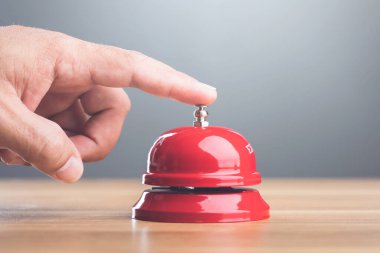 Gri arka planda duran ahşap bir masada kırmızı servis çanına el basarak. Görüntü acil ve harekete geçmeye hazır olduğunu iletir