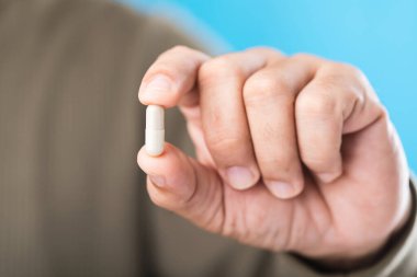 Mavi zemin üzerinde kapsül tutan bir kişinin yakın plan fotoğrafı, uygun ilaç kullanımının önemini vurgulayan ve her dozdan önce etiketleri dikkatle okuması gerektiğini hatırlatan