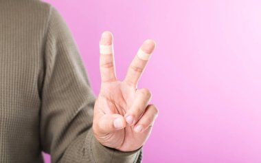 İndeks ve orta parmaklarda yara bandı olan V işareti oluşturan bir el, esnekliği ve küçük yaralanmalara rağmen çalışmaya devam edebilme yeteneğini sembolize ediyor.