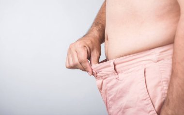 Daha büyük bir fiziğe sahip bir adam pantolonunun bel ölçüsünü düzelterek vücut şeklini ve genel sağlığını izlemenin önemini yansıtan sıkı mı gevşek mi diye kontrol eder.