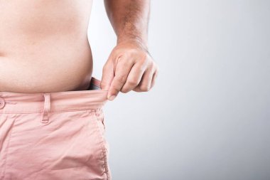 Daha büyük bir fiziğe sahip bir adam pantolonunun bel ölçüsünü düzelterek vücut şeklini ve genel sağlığını izlemenin önemini yansıtan sıkı mı gevşek mi diye kontrol eder.