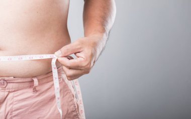 Daha büyük bir fiziği olan bir adam kemerini bir mezura ile ölçüyor ve sağlığına odaklanıyor. Bu, düzenli sağlık kontrollerinin ve erkekler için sağlıklı bir yaşam tarzının korunmasının önemini vurguluyor