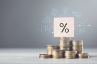 Tahta blok üzerinde '%' sembolü ve düzgünce istiflenmiş madeni paralar, yükselen faiz oranları konseptini sembolize ediyor, gri