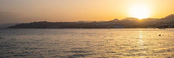Panoramic view of the sun setting behind the mountains of Eilat, Israel. People in silhouette enjoying the remaining daylight in the Red Sea.