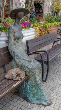 İsrail 'de bir bankta elbise ve şapka giymiş bir kadının bronz heykelinin yanında uyuyan başıboş gri bir tekir kedi..