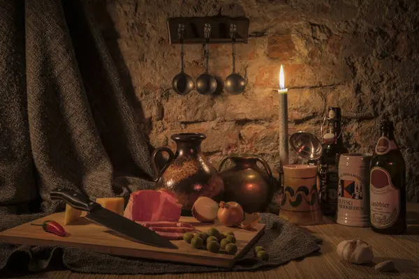 Masanın üzerinde iki bira bardağı, iki şişe bira, iki cuishins, masanın üzerinde bir parça et, zeytin, sarımsak, soğan, peynir ve bir biber, bir mum yanıyor.