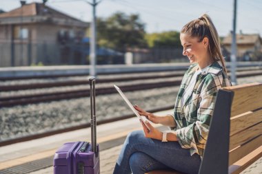 Tren istasyonunda trenini beklerken dijital tablet kullanan mutlu kadın..