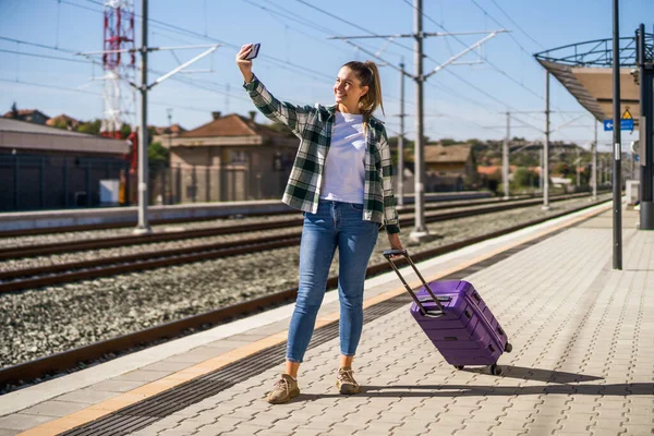 幸せな女は電車の駅に立っている間に携帯電話で自撮りしています — ストック写真