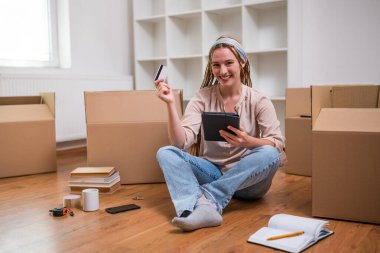 Örgülü modern kızıl kadın yeni evine taşınırken dijital tablet ve kredi kartı kullanıyor..