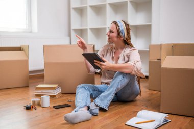 Yeni daireye taşınırken dijital tablet kullanan örgülü modern kızıl kadın..