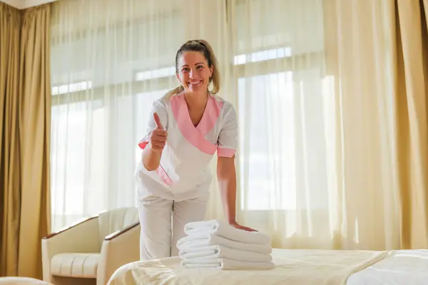 Beautiful hotel maid putting fresh clean towels on bed in room and showing thumb up.