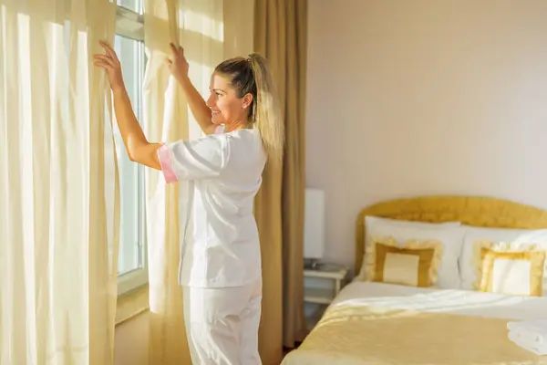 Image of beautiful  hotel maid opening window curtains  in a room.