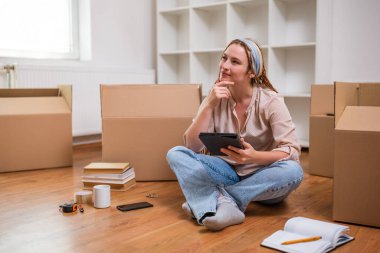 Yeni daireye taşınırken dijital tablet kullanan örgülü modern kızıl kadın..