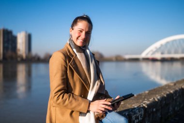 Sıcak giyimli güzel bir kadın dijital tablet kullanıyor ve güneşli bir kış gününde nehir kenarında dinlenmekten zevk alıyor..