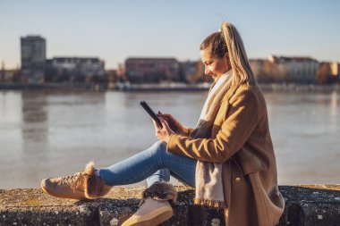 Dijital tablet kullanan ve güneşli bir kış gününde nehir kenarında dinlenmekten hoşlanan kalın giysili güzel bir kadın. Tonu ayarlanmış resim.