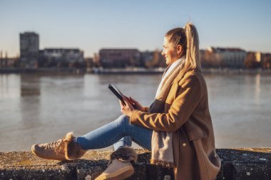 Dijital tablet kullanan ve güneşli bir kış gününde nehir kenarında dinlenmekten hoşlanan kalın giysili güzel bir kadın. Tonu ayarlanmış resim.
