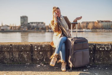 Bavullu mutlu bir turist nehir kenarında dinlenirken telefonla konuşuyor. Tonu ayarlanmış resim.