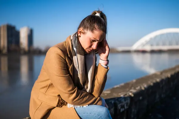 川のそばに座っている間に頭痛を抱えている心配な女性 — ストック写真