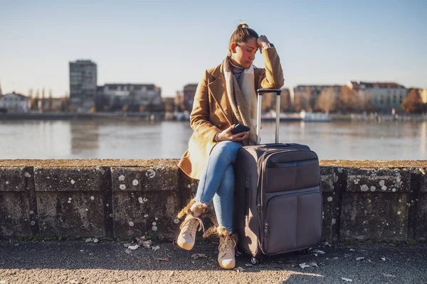 スーツケースと電話で心配した女性観光客は 川のそばに座って考える都市で迷子になった トーンされたイメージ — ストック写真