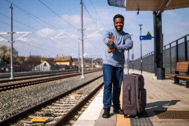 Bavullu mutlu adam tren istasyonunda dikiliyor ve başparmağını gösteriyor..