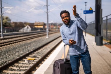 Elinde bavul ve cep telefonu olan kızgın bir adam tren istasyonunda bekliyor..