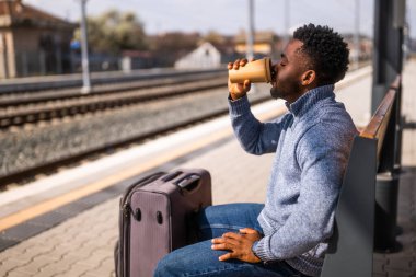 Bavullu mutlu adam tren istasyonunda bir bankta otururken kahve içmekten zevk alıyor..