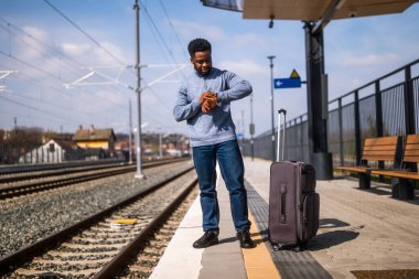 Endişeli bir adam, tren istasyonunda elinde bavulla saatine bakıyor..