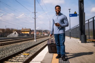 Tren istasyonunda yürürken telefonu kullanan bavullu mutlu adam..