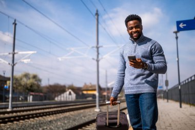 Tren istasyonunda beklerken telefonu kullanan bavullu mutlu bir adam..