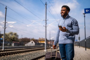 Tren istasyonunda beklerken telefonu kullanan bavullu mutlu bir adam..