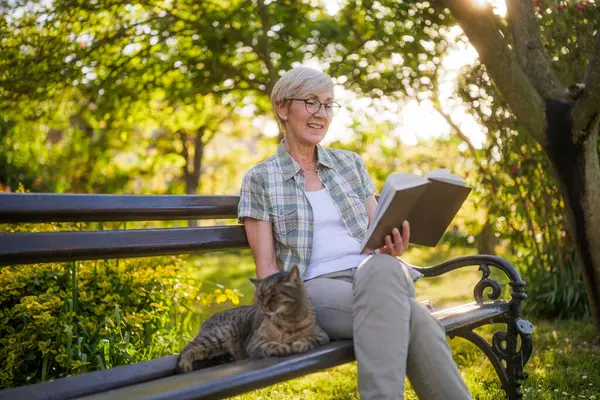 快乐的老妇人喜欢看书 喜欢在自家花园的长椅上和猫呆在一起 免版税图库图片