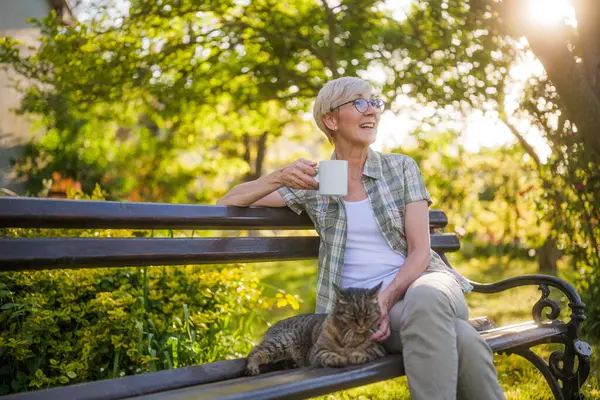 ハッピーシニアの女性は 彼女の庭のベンチに座っている間 コーヒーを飲んで彼女の猫と過ごす時間を楽しんでいます ストック写真
