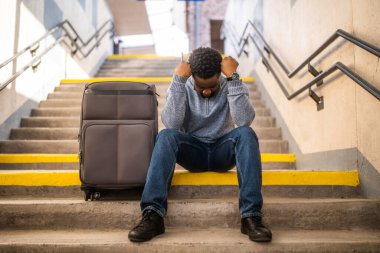 Tren istasyonunda merdivenlerde oturan telefonu ve bavulu olan endişeli bir adam..