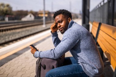 Elinde bavul ve cep telefonu olan kızgın bir adam tren istasyonundaki bankta oturuyor..