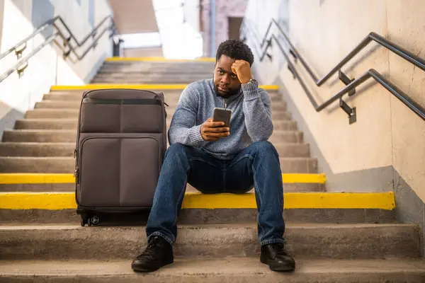 鉄道駅の階段に座っている電話とスーツケースを持つ心配した男 ストック画像