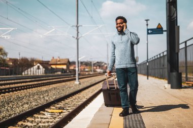 Bavullu mutlu adam tren istasyonunda yürürken telefonla konuşuyor..