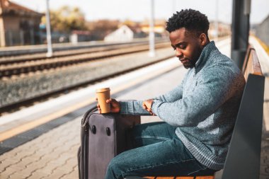 Elinde bavul ve kahve olan bir adam tren istasyonundaki bankta otururken saate bakıyor..