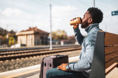 Bavullu bir adam tren istasyonunda bankta otururken kahve içmekten hoşlanır..