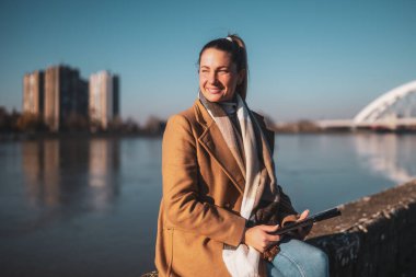 Sıcak giyimli güzel bir kadın dijital tablet kullanıyor ve güneşli bir kış gününde nehir kenarında dinlenmekten zevk alıyor..