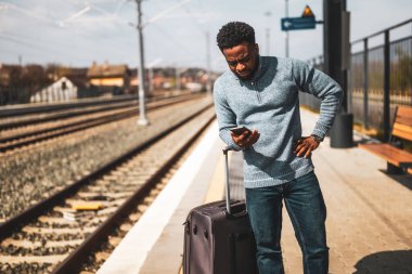 Tren istasyonunda duran bavulu ve cep telefonu olan endişeli bir adam..