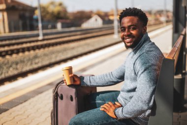 Bavullu mutlu adam tren istasyonunda bir bankta otururken kahve içmekten zevk alıyor..